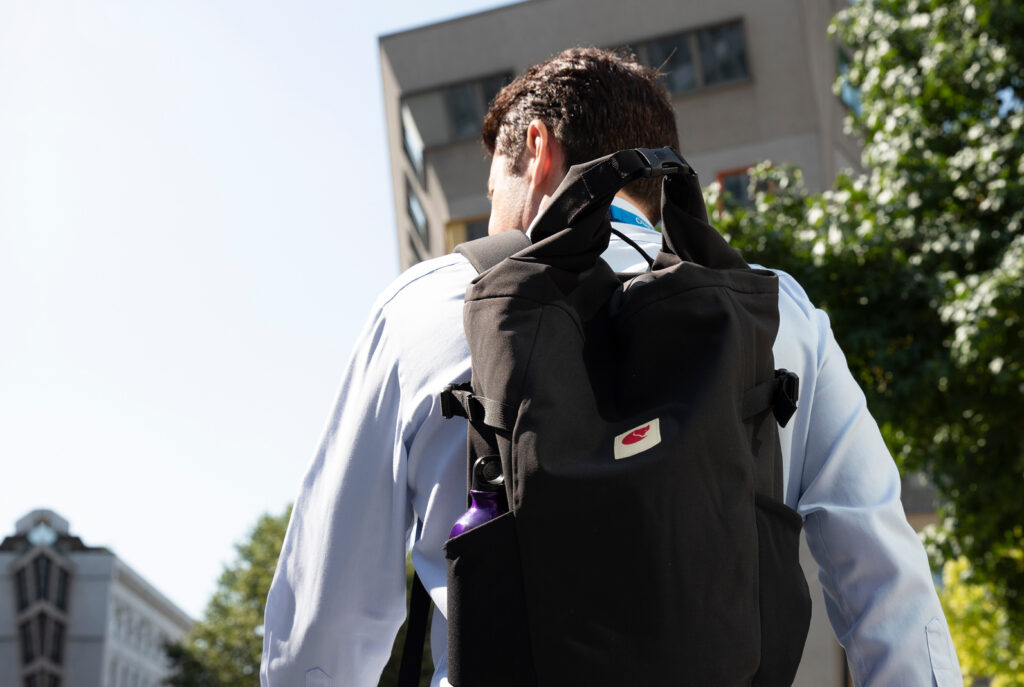 Image of a man with a backpack walking away.  He's in a city, there are office blocks in the background.  