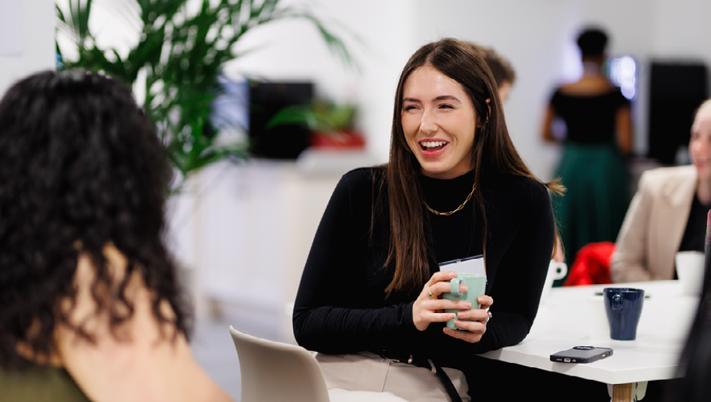 image of a person sitting holding a cup in a work environment, talking to another person